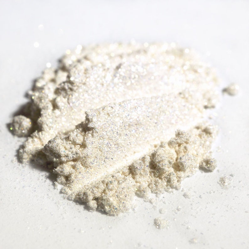 May include: Close-up of a pile of shimmering, pearlescent powder. The powder has a fine texture and reflects light, creating a sparkling effect. The color is a soft, off-white with hints of silver and gold. This product is likely a cosmetic, such as eyeshadow or highlighter.