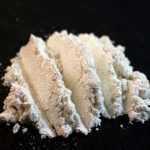 May include: Close-up of a pile of pearlescent powder with a shimmering, glittery effect. The powder is a light, creamy color with visible sparkle, creating a luminous appearance. The background is a solid black.