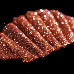 May include: A close-up of a pile of copper glitter with a shimmery, iridescent finish. The glitter is arranged in a wave-like pattern, creating a textured and sparkling effect.