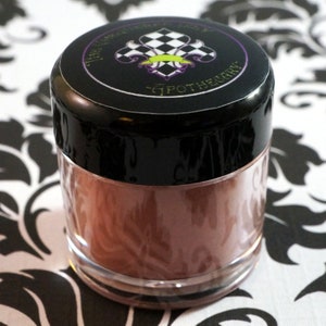 May include: A small, clear jar with a black lid. The lid has a black and white checkered design with a green and purple fleur-de-lis. The jar contains a brown powder.