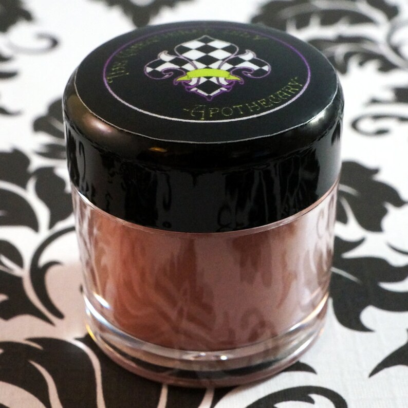May include: A small, clear jar with a black lid. The lid has a black and white checkered design with a green and purple fleur-de-lis. The jar contains a brown powder.