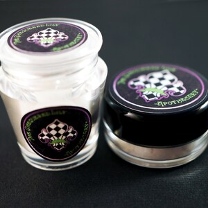 Pu&ograve; includere: Due piccoli barattoli di vetro con coperchi ed etichette neri. Le etichette sono nere con un design a scacchi bianco e nero e un giglio verde e viola. Il testo sull'etichetta recita "The Green Lily Apothecary".
