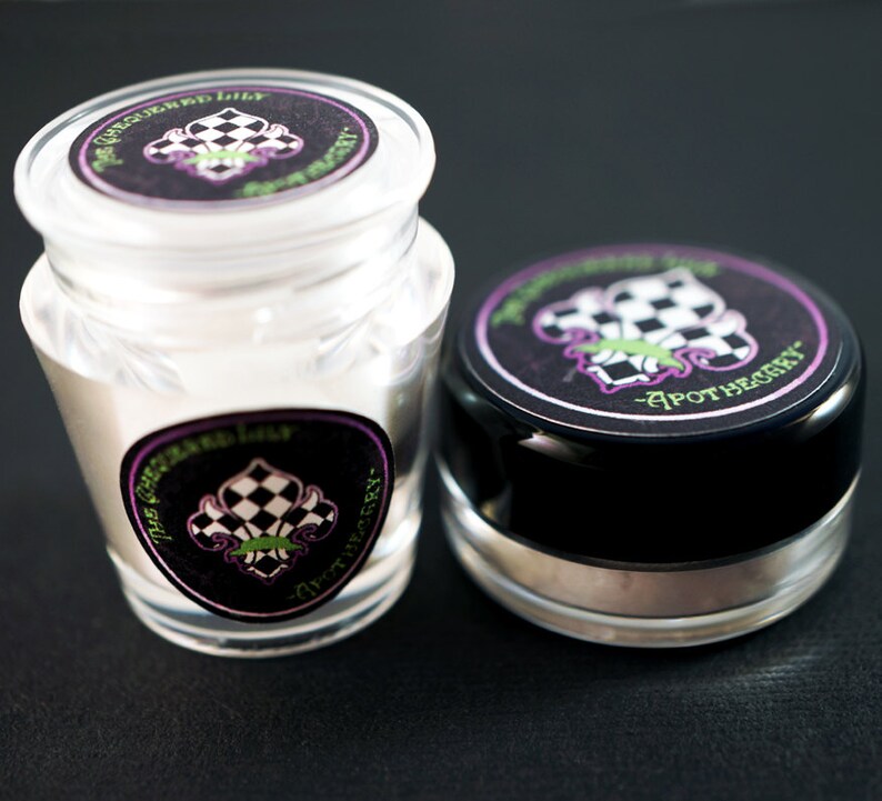 Pu&ograve; includere: Due piccoli barattoli di vetro con coperchi ed etichette neri. Le etichette sono nere con un design a scacchi bianco e nero e un giglio verde e viola. Il testo sull'etichetta recita "The Green Lily Apothecary".