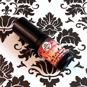 Op de afbeelding: Een klein, zwart flesje met een etiket waarop "Bubble Gum" en "Sugar, Sweetness, & Paradise" staat in een roze, oranje en gele gradiënt. Het flesje staat op een wit en zwart gepatroneerd oppervlak.