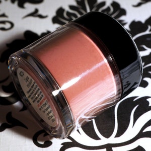 May include: A clear jar with a black lid filled with a light pink powder. The jar has a label with the text "The  L.A. Girl  Pro  Cosmetics  Blush  The  Blush  is  a  highly  pigmented  powder  blush  that  gives  you  a  natural  flush  of  color  Net  Wt  1.5g  (0.05  oz)"