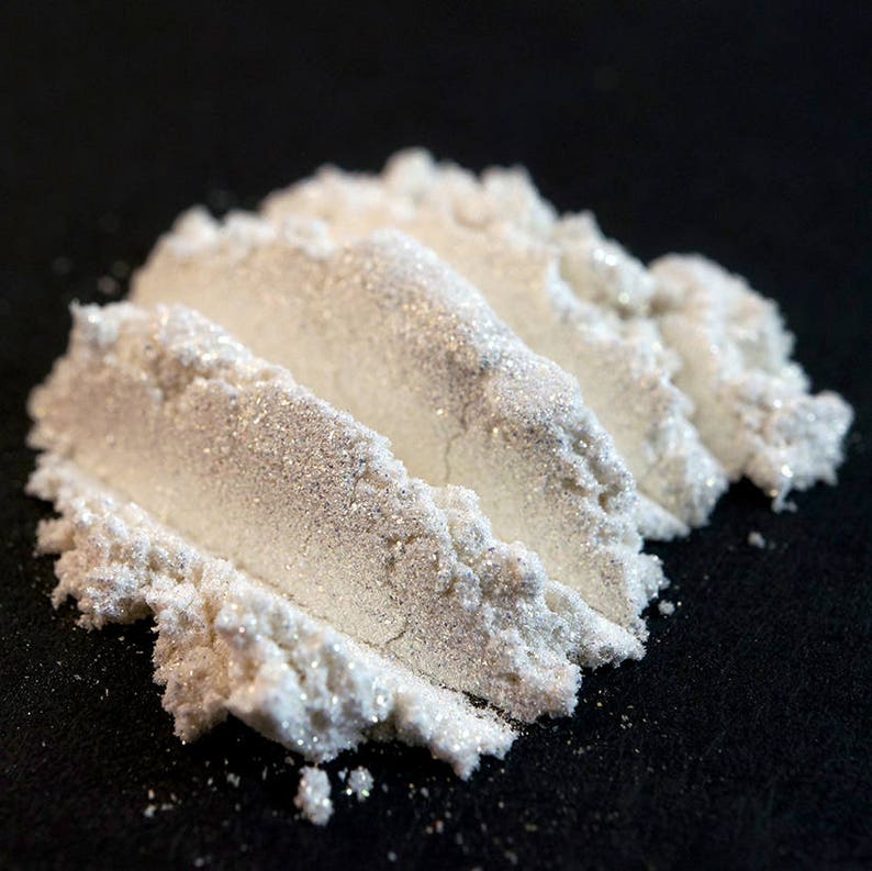 May include: Close-up of a pile of white, shimmering powder with a pearlescent effect. The powder is arranged in three parallel lines on a black surface, creating a textured appearance. The fine particles catch the light, giving a sparkly look.