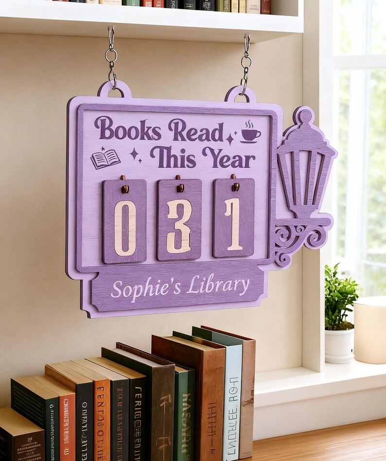Peut inclure: Panneau en bois lilas affichant "Books Read This Year" et le nombre "031". Le panneau pr&eacute;sente une lanterne d&eacute;corative et est personnalis&eacute; avec "Sophie's Library". Il est suspendu par des cha&icirc;nes au-dessus d'une &eacute;tag&egrave;re avec des livres.