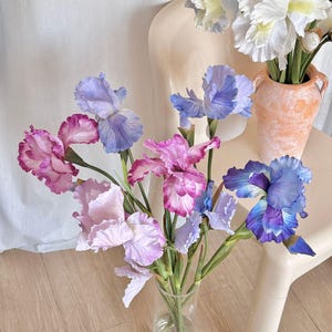May include: A bouquet of artificial iris flowers in a clear glass vase. The irises display a range of colors, including shades of purple, blue, and pink. The flowers are arranged with green stems, creating a vibrant floral display.