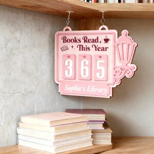 Peut inclure: Panneau en bois rose avec l'inscription "Books Read This Year" et le chiffre "365". Le panneau, suspendu par des cha&icirc;nes, pr&eacute;sente une lanterne d&eacute;corative. Une pile de livres est visible en dessous, avec le texte "Sophie's Library".