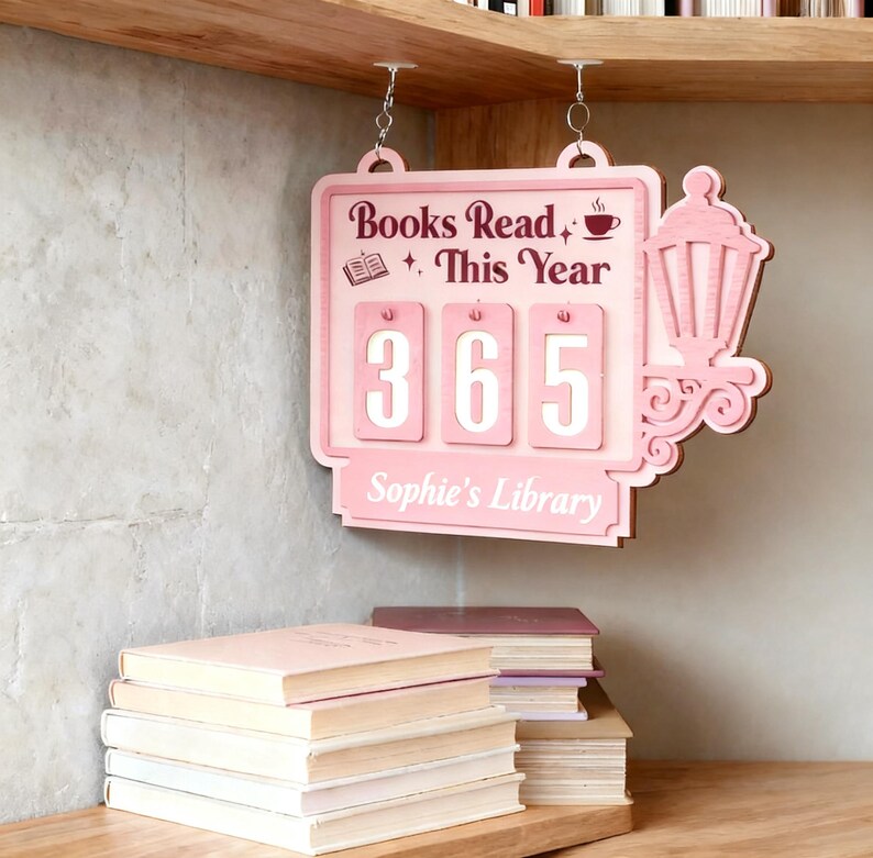 Peut inclure: Panneau en bois rose avec l'inscription "Books Read This Year" et le chiffre "365". Le panneau, suspendu par des cha&icirc;nes, pr&eacute;sente une lanterne d&eacute;corative. Une pile de livres est visible en dessous, avec le texte "Sophie's Library".