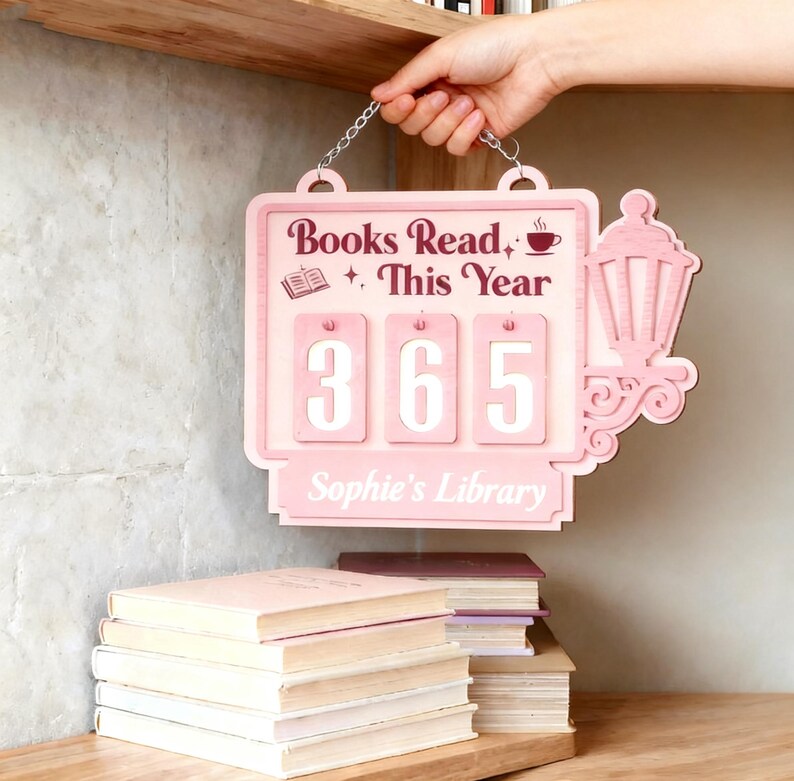 Peut inclure: Panneau en bois rose avec l'inscription "Books Read This Year" et le chiffre "365". Le panneau est orn&eacute; d'une lanterne d&eacute;corative et est suspendu &agrave; une cha&icirc;ne. Le nom "Sophie's Library" est &eacute;galement pr&eacute;sent. Une pile de livres est visible.
