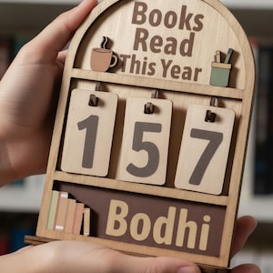 Può includere: Contatore di libri in legno con la scritta "Books Read This Year" e il numero "157". Presenta icone di tazza di caffè e portapenne. Il nome "Bodhi" è in basso. Un regalo unico per gli amanti della lettura.