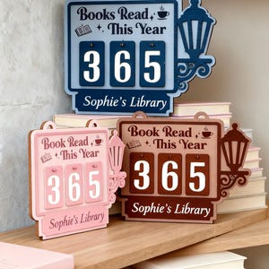 Peut inclure: Trois compteurs de livres en bois bleus, roses et marron, affichant chacun "Livres lus cette ann&eacute;e" et le nombre "365". Les compteurs ont un motif de lanterne et le texte "Sophie's Library".