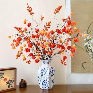 Könnte beinhalten: Ein Blumenarrangement aus orangefarbenen und roten Blüten in einer Vase mit weißem und blauem Muster. Die Vase steht auf einer Holzoberfläche, mit einem gerahmten Kunstwerk und einer kleinen Holzskulptur. Die Blüten haben eine einzigartige, laternenartige Form.