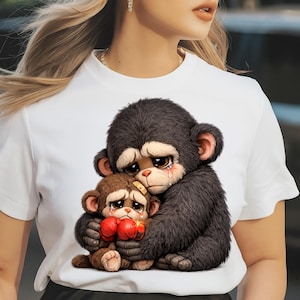 May include: White t-shirt featuring a graphic of two sad cartoon monkeys hugging. The larger monkey is dark brown with a bandage on its head, and the smaller monkey is brown and wearing red boxing gloves. Both have tears.