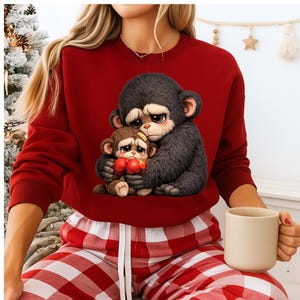 May include: A red sweatshirt featuring a graphic of two sad cartoon monkeys hugging. The larger monkey is dark gray, and the smaller monkey is brown, holding red boxing gloves. The monkeys have teary eyes.