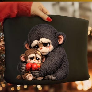 May include: A black pillow featuring a digital illustration of two sad cartoon monkeys hugging. The larger monkey has dark gray fur, and the smaller monkey has brown fur and red boxing gloves. Both monkeys have tears.