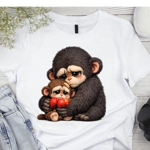 May include: White t-shirt featuring a graphic of two sad cartoon monkeys hugging. The larger monkey is dark brown with a tear rolling down its cheek. The smaller monkey is brown and wearing red boxing gloves.