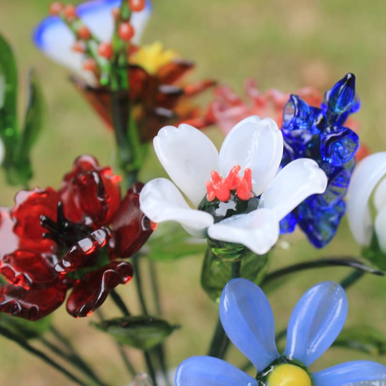 May include: A close-up of a bouquet of colorful glass flowers. The flowers include red, white, blue, and yellow petals with green stems. The glass flowers are arranged in a natural setting with a blurred green background.