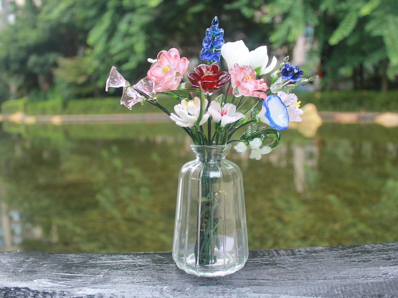 May include: A clear glass vase filled with colorful glass flowers. The flowers include pink, red, white, and blue blooms. The vase is sitting on a wooden surface with a blurred green background.