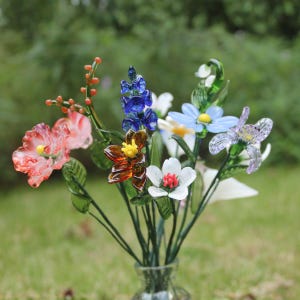 May include: A bouquet of colorful glass flowers in a clear glass vase. The flowers include pink, blue, white, and orange blooms with green stems and leaves. The background is a blurred green.