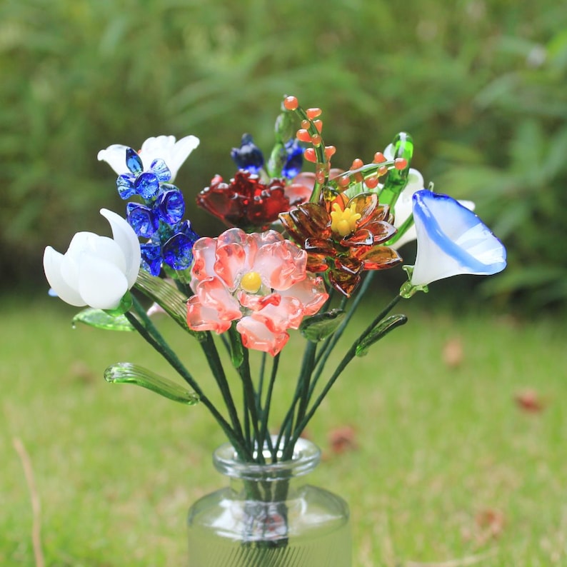May include: A colorful bouquet of glass flowers in a clear glass vase. The arrangement includes white, blue, red, and orange flowers with green stems. The vase sits on a bed of green grass.