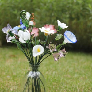 May include: A clear glass vase holds a colorful bouquet of glass flowers. The arrangement includes white calla lilies, pink and white daisies, blue morning glories, and star-shaped accents. The flowers are set against a blurred green background.