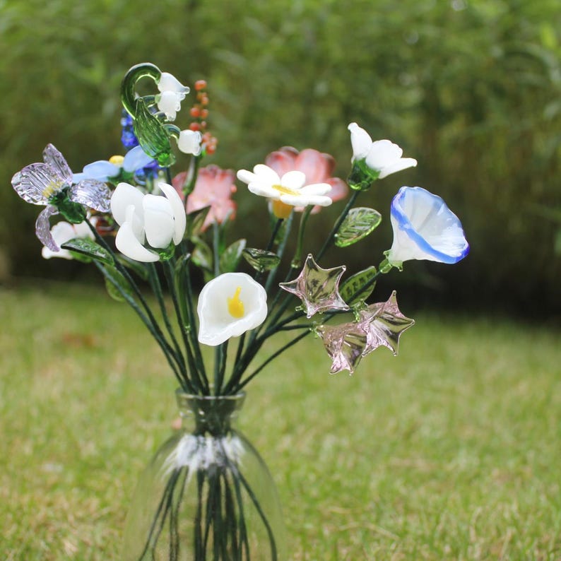 May include: A clear glass vase holds a colorful bouquet of glass flowers. The arrangement includes white calla lilies, pink and white daisies, blue morning glories, and star-shaped accents. The flowers are set against a blurred green background.