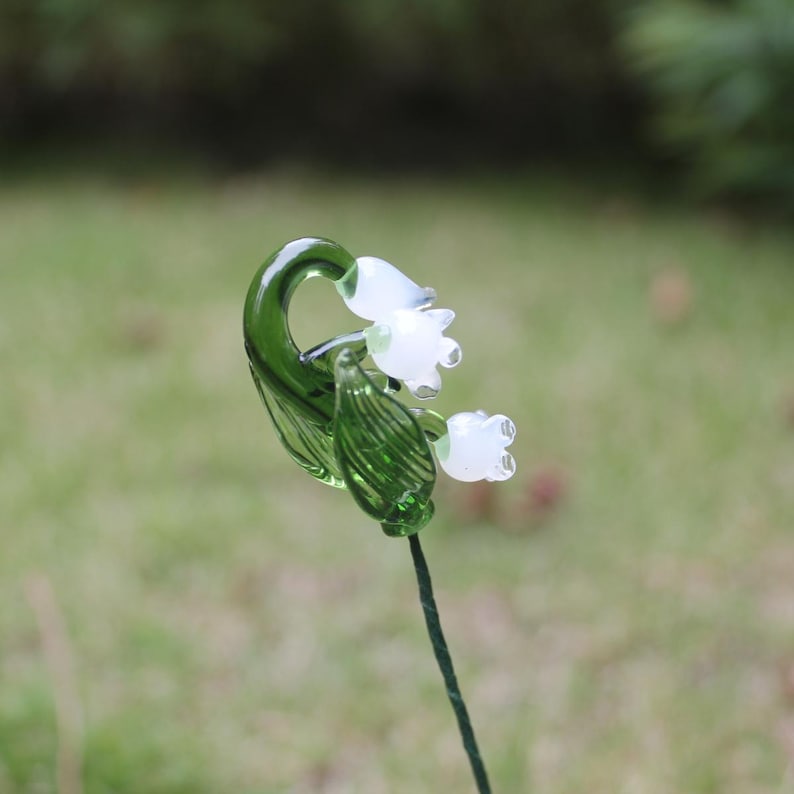 May include: A glass lily of the valley flower with white blossoms and green leaves and stem. The flower is a delicate piece of art, perfect for garden decor or as a unique gift. The glass is clear and reflects light.