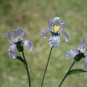 May include: Three glass iris flowers with purple petals and yellow centres. The stems are dark green, and the flowers are set against a blurred green background. The glass has a textured, crystalline appearance.