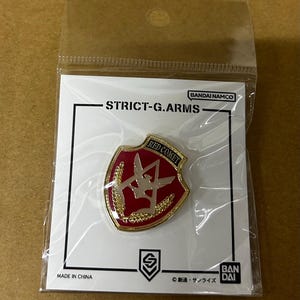 Könnte beinhalten: Ein roter und goldener Emaille-Pin mit dem Text "RED COMET" und "STRICT-G.ARMS" auf einer weißen Karte. Der Pin ist schildförmig mit einem goldenen Rand und einem stilisierten Design. Der Pin befindet sich in einer transparenten Kunststoffverpackung.