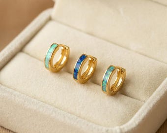 May include: Three gold-colored hoop earrings with opal inlays in shades of blue and green are displayed in a beige jewelry box. The earrings are small and have a polished finish, showcasing the iridescent quality of the opal.