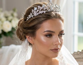 May include: A silver-toned bridal tiara with clear crystals and floral accents. The tiara features a central, larger crystal and smaller crystals throughout the design. The image shows a woman wearing the tiara with a veil.