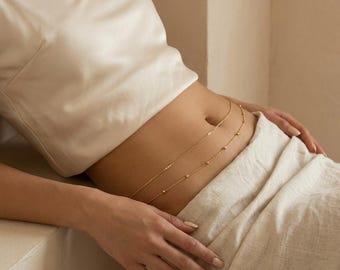 May include: A delicate gold-colored body chain with small, evenly spaced beads. The chain is draped across the waist, creating a subtle, elegant accessory. The model is wearing a white satin top and light-colored pants.