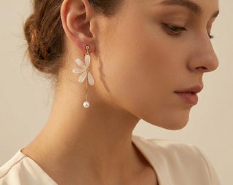 May include: A gold-toned earring featuring a white flower-shaped charm with five petals, suspended from a thin gold wire that ends in a small white pearl bead. The earring is designed as a clip-on style, showcasing a delicate and slender aesthetic.