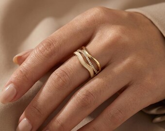 May include: A gold-toned ring set with three bands. One band is white enamel, another is a wave design, and the third is encrusted with small, clear gemstones. The ring is on a person's finger.