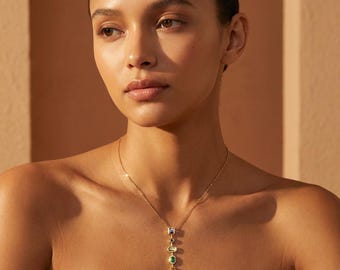 May include: A gold necklace with a series of colorful gemstones, including green, red, and blue, hangs against a warm-toned background. The necklace is the focal point, with the gemstones arranged in a vertical line. The model's skin tone is a warm brown.