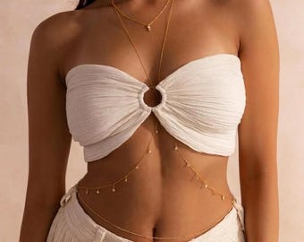 May include: A gold-colored body chain with a delicate design. The chain features multiple strands that drape across the chest and waist, adorned with small, sparkling accents. The jewelry is worn over a white, textured top and pants.
