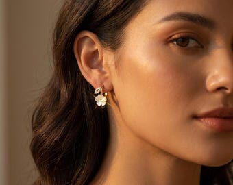 May include: Gold-tone hoop earrings with a floral design. Each earring features a small white flower and a leaf-shaped accent with clear gemstones. The earrings are shown on a person's ear.