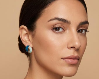 May include: A close-up of a person wearing a pair of blue and light blue hoop earrings. The earrings are a half-circle shape and have a glossy finish. The background is a solid, neutral color.