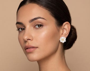 May include: White flower-shaped stud earrings with a gold center. The earrings are worn on the ear of a person with dark hair and a neutral skin tone. The background is a solid, light beige color.