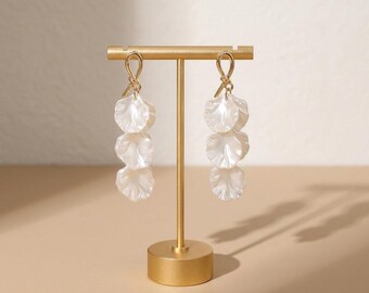 May include: A pair of gold-toned drop earrings featuring a knotted top design and three stacked, translucent white petal-shaped components. The earrings are displayed on a gold-colored T-shaped metal stand with a circular base.