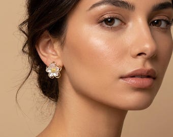 May include: A close-up of a flower-shaped earring. The earring features a gold-colored frame with a white flower and a yellow center. The earring is worn on a person's ear.