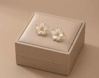 May include: A pair of flower-shaped stud earrings. Each earring features a white, petal-like design with a gold-colored outline and a yellow center. The earrings are displayed on a beige jewelry box.