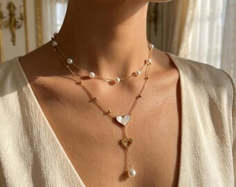May include: A gold-toned necklace with two strands, adorned with pearls and small gold beads. The lower strand features a heart-shaped pendant with a mother-of-pearl inlay and a dangling pearl, creating an elegant and feminine accessory.