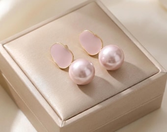 May include: A pair of elegant earrings featuring a light pink, teardrop-shaped design with a gold trim, paired with a large, round, pearl-like bead. The earrings are displayed on a beige jewelry box, creating a sophisticated presentation.
