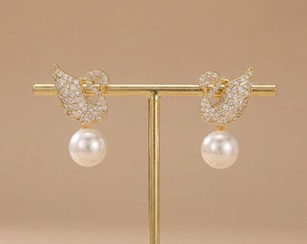 May include: A pair of gold-toned swan earrings. Each earring features a swan design encrusted with small, clear crystals and a large, white, pearlescent drop. The earrings are displayed on a gold-colored stand against a neutral background.