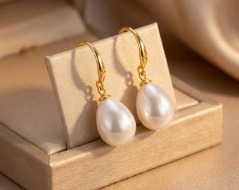 May include: A pair of gold-colored drop earrings with pear-shaped, white pearls. The earrings are displayed in a tan jewelry box, with a soft, blurred background.
