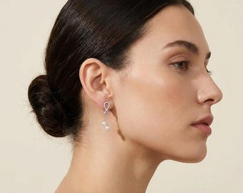 May include: A silver-toned drop earring featuring a small, sparkling knot-shaped stud at the top, connected to a delicate chain that suspends two small, round, white pearls. The earring is worn on the earlobe, showcasing a refined, dangling design.
