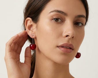 May include: Cherry-shaped earrings with a gold-colored post and a deep red, glossy finish. The earrings are dangling, with a cherry design. The earrings are a fun and playful accessory.